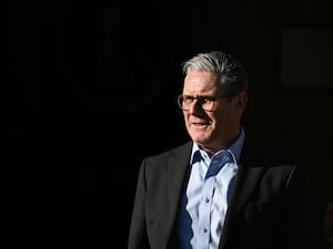 Sir Keir Starmer in a dark suit jacket and light blue open-necked shirt against a black background