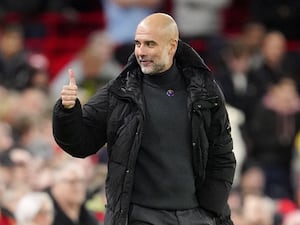 Manchester City manager Pep Guardiola, wearing a black coat, gives someone the thumbs-up while on the touchline