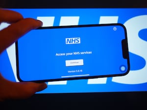 A smartphone displaying the NHS app in front of an NHS sign