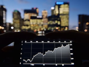 A person views share prices on an Iphone, with London’s Canary Wharf in the background