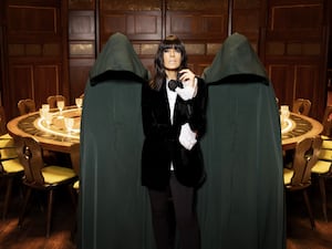 Claudia Winkleman flanked by two people dressed in hooded gowns, with the hoods up hiding their faces