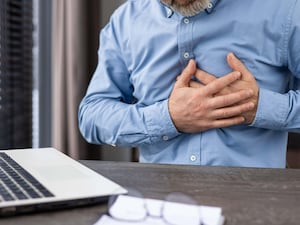 A middle-aged man in a blue shirt clutches his chest in pain while working on a laptop in a professional office setting. The image conveys stress, discomfort, and potential heart health issues.