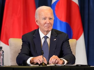 Joe Biden smiles, while sat at a desk before national flags