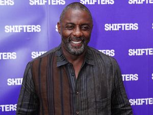 Idris Elba, wearing a brown and grey striped shirt, smiling in front of a purple backdrop featuring the word Shifters repeatedly in white at the Shifters West End premiere after-show party in London