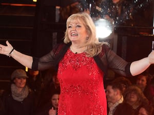 Linda Nolan in a red dress and black shawl holding her arm out in celebration