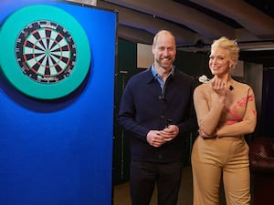 Prince William and Hannah Waddingham holding darts and standing next to a dart board mounted on a blue wall