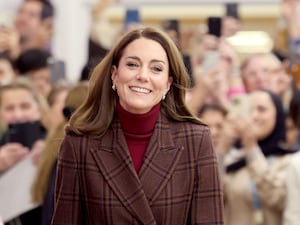 Princess of Wales walks alongside a crowd, she is in a checked brown coat, red turtleneck jumper and she is smiling