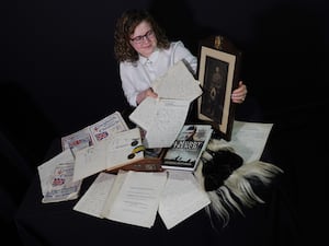 Woman holding old-looking sheets of paper with hand writing on and a black and white photo of a soldier in a kilt, with other papers and a sporran spread out in front of her