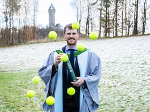 Jamie Murray holding a tube and wearing a graduation gown, with tennis balls thrown in the air in front of him