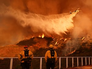 Two firefighters with their back to the camera watch as a plane dumps water on a burning landscape