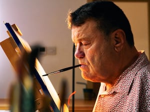 A man paints on a canvas using a paintbrush held between his lips